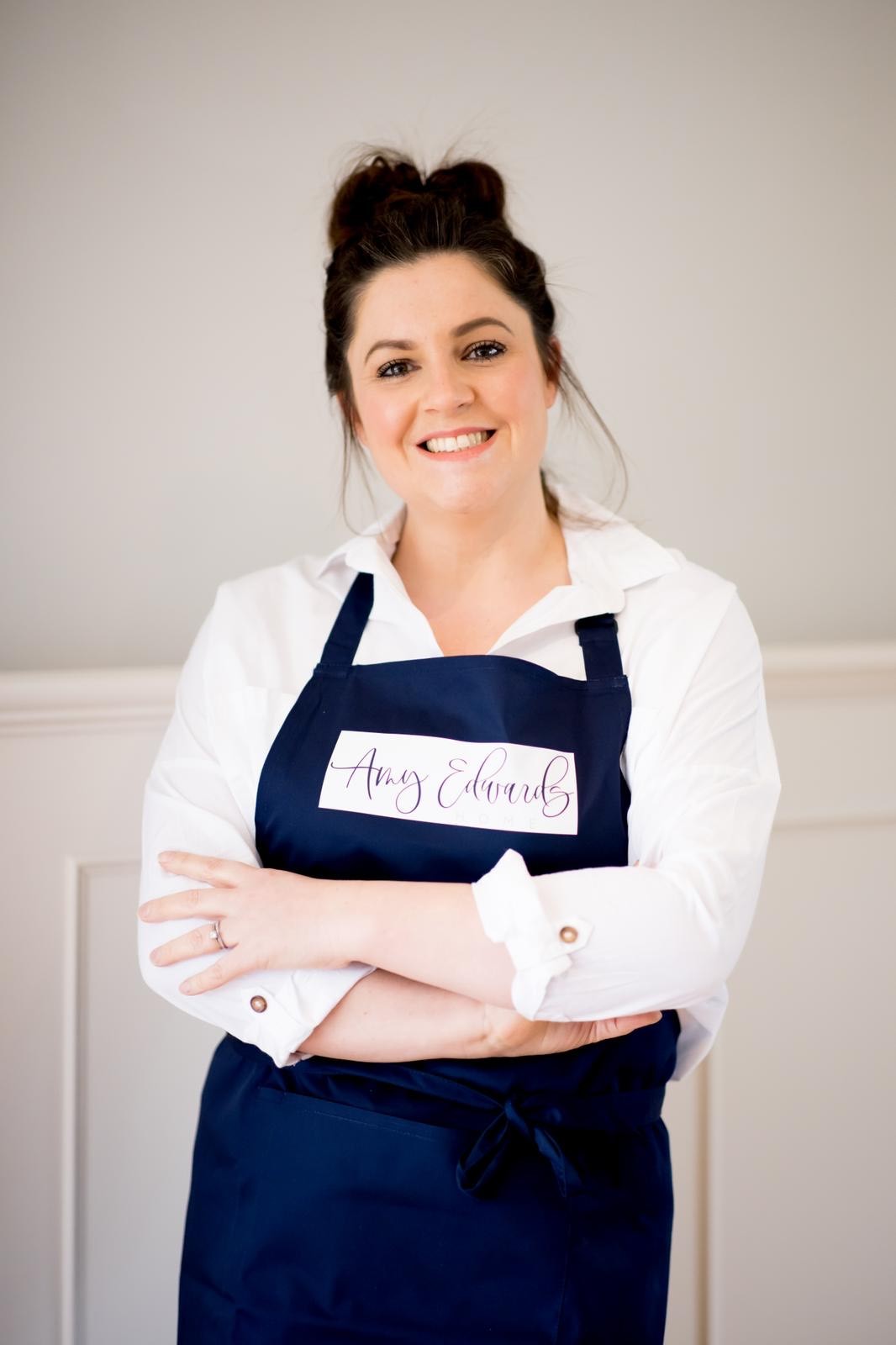 Amy Edwards cross armed smiling at the camera. Wearing a white shirt and navy painting apron.